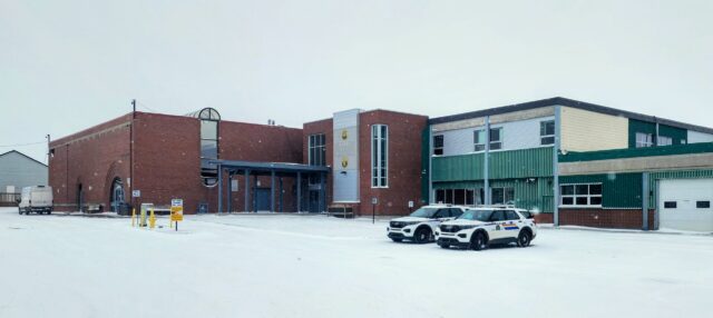 A brick and green municipal building in a snowy parking lot with two police SUVs parked in front.