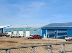 New Overhead Doors & LED Lighting Approved For Town Municipal Shop Blue industrial warehouse complex with multiple loading bays and several pickup trucks parked on a dirt lot, enclosed by a chain-link fence under a clear blue sky.