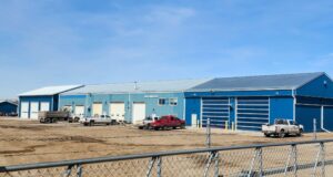 New Overhead Doors & LED Lighting Approved For Town Municipal Shop Blue industrial warehouse complex with multiple loading bays and several pickup trucks parked on a dirt lot, enclosed by a chain-link fence under a clear blue sky.