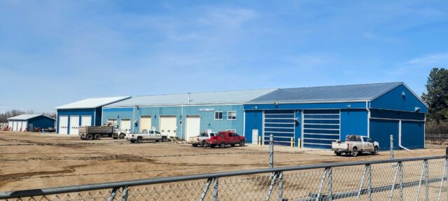 Blue industrial warehouse complex with multiple loading bays and several pickup trucks parked on a dirt lot, enclosed by a chain-link fence under a clear blue sky.