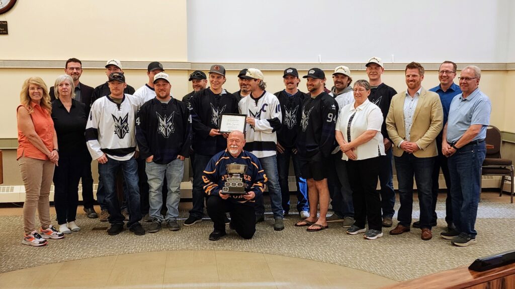 Group of people posing in a hall; a man kneels in front holding a trophy while teammates in hockey jerseys stand behind with a certificate center stage.