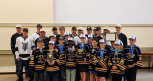 Stettler Hockey Teams Invited To Town Council To Celebrate Championship Wins Group of young hockey players in navy and gold jerseys with medals, posing for a team photo with a large trophy and certificate; coaches stand behind them in an indoor venue.