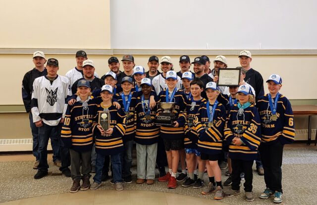 Group of young hockey players in navy and gold jerseys with medals, posing for a team photo with a large trophy and certificate; coaches stand behind them in an indoor venue.