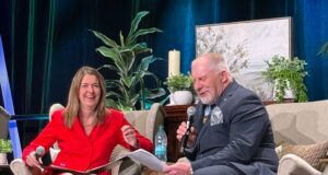 Stettler County Councilor Hosts Premier at ASCHA Housing Convention Two people sit on stage in beige armchairs, talking into handheld microphones; woman in red dress smiles as she gestures with papers in lap, man in dark suit reads from a sheet.