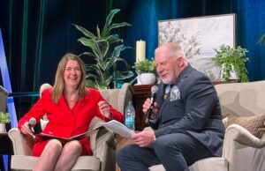 Stettler County Councilor Hosts Premier at ASCHA Housing Convention Two people sit on stage in beige armchairs, talking into handheld microphones; woman in red dress smiles as she gestures with papers in lap, man in dark suit reads from a sheet.