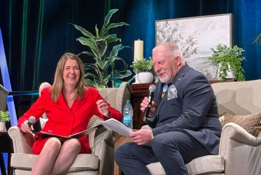 Stettler County Councilor Hosts Premier at ASCHA Housing Convention Two people sit on stage in beige armchairs, talking into handheld microphones; woman in red dress smiles as she gestures with papers in lap, man in dark suit reads from a sheet.