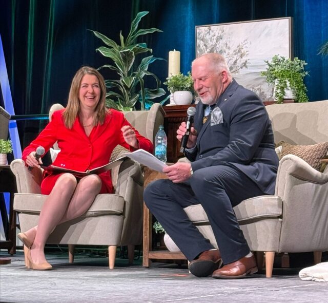 Two people sit on stage in beige armchairs, talking into handheld microphones; woman in red dress smiles as she gestures with papers in lap, man in dark suit reads from a sheet.