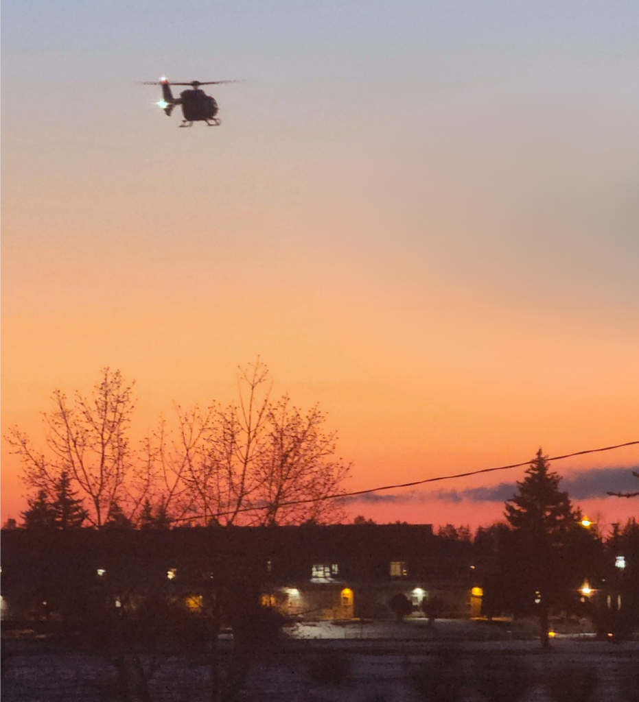 Silhouette of a helicopter flying over a tree-lined urban area at sunset with streetlights glowing below.