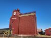 Future of Stettler P&H Grain Elevator Uncertain Following Contentious AGM Red grain elevator building with STETTLER lettering and a PH shield logo, set against a bright blue sky; a vintage green Prairie train car sits in front behind a fence.