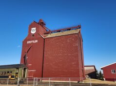 Future of Stettler P&H Grain Elevator Uncertain Following Contentious AGM Red grain elevator building with STETTLER lettering and a PH shield logo, set against a bright blue sky; a vintage green Prairie train car sits in front behind a fence.