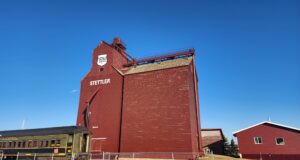 Future of Stettler P&H Grain Elevator Uncertain Following Contentious AGM Red grain elevator building with STETTLER lettering and a PH shield logo, set against a bright blue sky; a vintage green Prairie train car sits in front behind a fence.