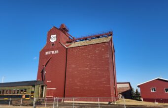 Future of Stettler P&H Grain Elevator Uncertain Following Contentious AGM Red grain elevator building with STETTLER lettering and a PH shield logo, set against a bright blue sky; a vintage green Prairie train car sits in front behind a fence.