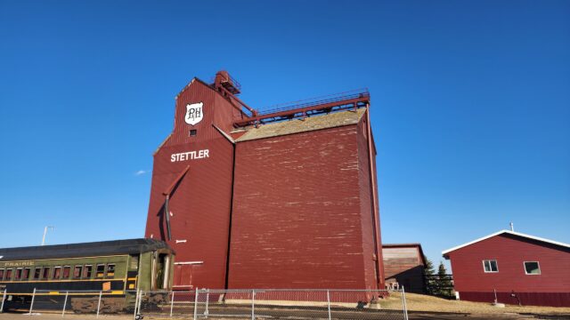 Red grain elevator building with STETTLER lettering and a PH shield logo, set against a bright blue sky; a vintage green Prairie train car sits in front behind a fence.