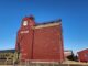 Future of Stettler P&H Grain Elevator Uncertain Following Contentious AGM Red grain elevator building with STETTLER lettering and a PH shield logo, set against a bright blue sky; a vintage green Prairie train car sits in front behind a fence.
