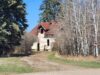 Stettler House Fire Deemed Suspicious; No Injuries Reported Abandoned beige house with a red roof partially hidden by bare trees along a dirt path under a clear blue sky.