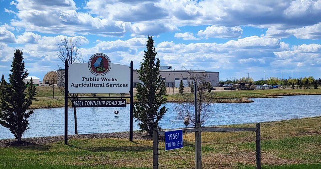 Public Works Agricultural Services sign with county seal beside a pond and a modern building in the background, 19561 Township Road 38-4 (TWP RD 38-4).