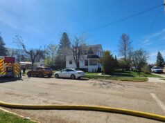 Stettler Fire Department Responds To House Fire Across From School Complex Firefighters connect a large hose to a fire truck in a suburban intersection on a sunny day, with a white house nearby.