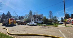 Stettler Fire Department Responds To House Fire Across From School Complex Firefighters connect a large hose to a fire truck in a suburban intersection on a sunny day, with a white house nearby.