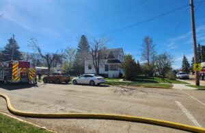 Stettler Fire Department Responds To House Fire Across From School Complex Firefighters connect a large hose to a fire truck in a suburban intersection on a sunny day, with a white house nearby.