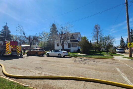 Stettler Fire Department Responds To House Fire Across From School Complex Firefighters connect a large hose to a fire truck in a suburban intersection on a sunny day, with a white house nearby.