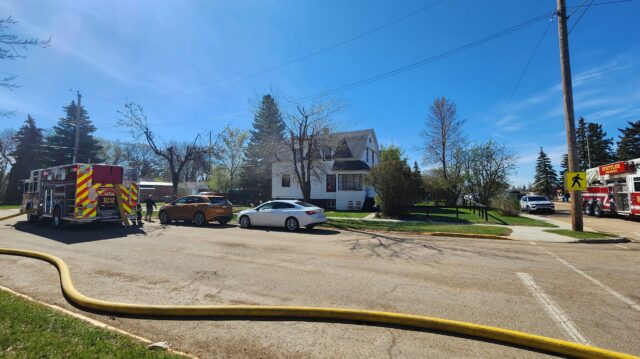 20260519_101737 Firefighters connect a large hose to a fire truck in a suburban intersection on a sunny day, with a white house nearby.