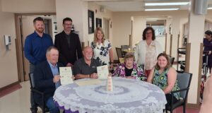 Stettler Town and County Partner for 40th Annual Seniors Week Celebrations Group of eight adults posing around a round table, some holding certificates and smiling in a bright, indoor room.
