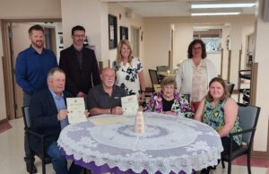 Stettler Town and County Partner for 40th Annual Seniors Week Celebrations Group of eight adults posing around a round table, some holding certificates and smiling in a bright, indoor room.