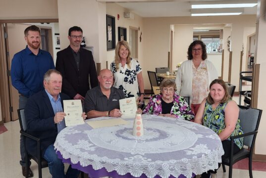 Stettler Town and County Partner for 40th Annual Seniors Week Celebrations Group of eight adults posing around a round table, some holding certificates and smiling in a bright, indoor room.