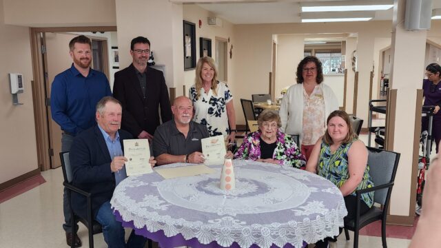20260519_112534 Group of eight adults posing around a round table, some holding certificates and smiling in a bright, indoor room.