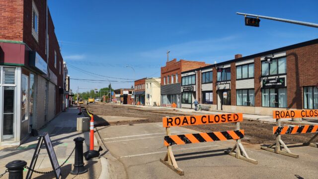 Street under construction with orange barriers reading 'ROAD CLOSED' and a closed curbside area on a sunny day in a commercial district.