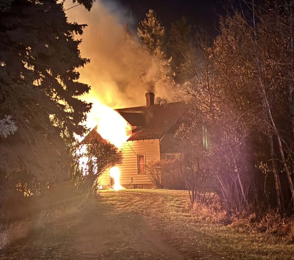 Nighttime scene of a house on fire with bright orange flames and dark smoke, trees framing the burning building and a chimney visible on the roof.