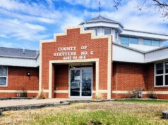Stettler County Scales Back Buffalo Lake Upgrade Plans For 2026 Brick county office building with a stepped brick facade and glass entrance doors, blue sky above, and a small grassy area in front.