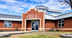 Stettler County Scales Back Buffalo Lake Upgrade Plans For 2026 Brick county office building with a stepped brick facade and glass entrance doors, blue sky above, and a small grassy area in front.