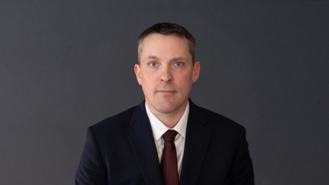 Nate Horner Professional headshot of a man in a navy suit and burgundy tie against a dark gray background.