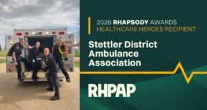 Stettler District Ambulance Association Awarded 2026 Rhapsody Healthcare Heroes Award Five paramedics pose in front of an open ambulance, celebrating a healthcare heroes award ceremony on a sunny day.