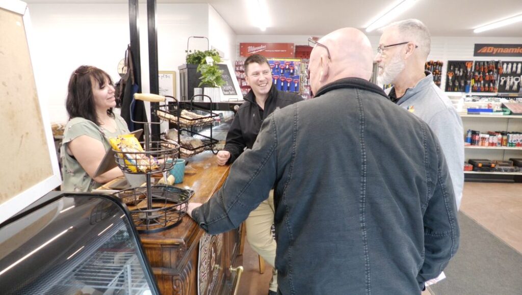 Clerk serving a customer at a wooden counter with takeaway baskets, while two men wait in a shop aisle in the background.