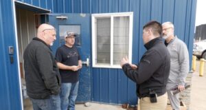 SREMA & Board of Trade Partner To Mobilize Local Business For Disaster Response Four men stand outside a blue metal building, casually chatting near a window and door.
