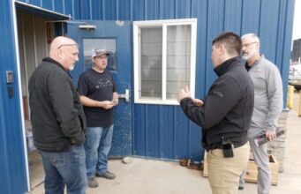 SREMA & Board of Trade Partner To Mobilize Local Business For Disaster Response Four men stand outside a blue metal building, casually chatting near a window and door.