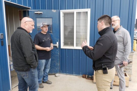 SREMA & Board of Trade Partner To Mobilize Local Business For Disaster Response Four men stand outside a blue metal building, casually chatting near a window and door.