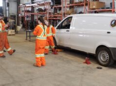 Stettler Young Women in Trades Workshop Gives Students Hands-On Experience Group of workers in high-visibility orange suits standing around a white van on jack stands in a workshop fallow area with shelves in the background.