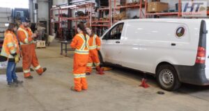 Stettler Young Women in Trades Workshop Gives Students Hands-On Experience Group of workers in high-visibility orange suits standing around a white van on jack stands in a workshop fallow area with shelves in the background.