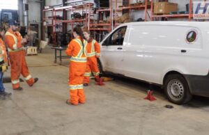 Stettler Young Women in Trades Workshop Gives Students Hands-On Experience Group of workers in high-visibility orange suits standing around a white van on jack stands in a workshop fallow area with shelves in the background.