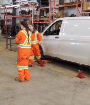 Stettler Young Women in Trades Workshop Gives Students Hands-On Experience Group of workers in high-visibility orange suits standing around a white van on jack stands in a workshop fallow area with shelves in the background.
