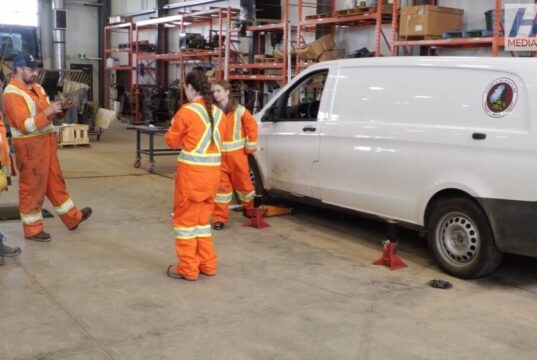 Stettler Young Women in Trades Workshop Gives Students Hands-On Experience Group of workers in high-visibility orange suits standing around a white van on jack stands in a workshop fallow area with shelves in the background.