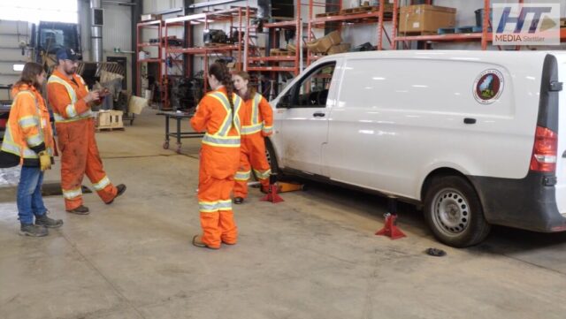 Screenshot 2026-05-21 135727 Group of workers in high-visibility orange suits standing around a white van on jack stands in a workshop fallow area with shelves in the background.