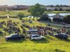 Stettler Town Council Approves July 1 Pancake Breakfast Outdoor park concert: a large crowd sits on the grass facing a small stage by the water, with Canadian flags on the stage pavilion and cars parked nearby.