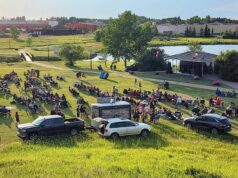 Stettler Town Council Approves July 1 Pancake Breakfast Outdoor park concert: a large crowd sits on the grass facing a small stage by the water, with Canadian flags on the stage pavilion and cars parked nearby.