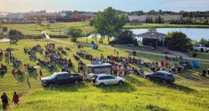 Stettler Town Council Approves July 1 Pancake Breakfast Outdoor park concert: a large crowd sits on the grass facing a small stage by the water, with Canadian flags on the stage pavilion and cars parked nearby.