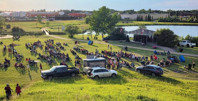 20250701_203140-EDIT Outdoor park concert: a large crowd sits on the grass facing a small stage by the water, with Canadian flags on the stage pavilion and cars parked nearby.