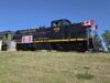 2026 Kin Canada ‘Ride the Rails’ Convention Held in Stettler Black steam locomotive with Canadian flags on the cabin, Alberta Prairie 1118, on a grassy rail yard under a clear blue sky.